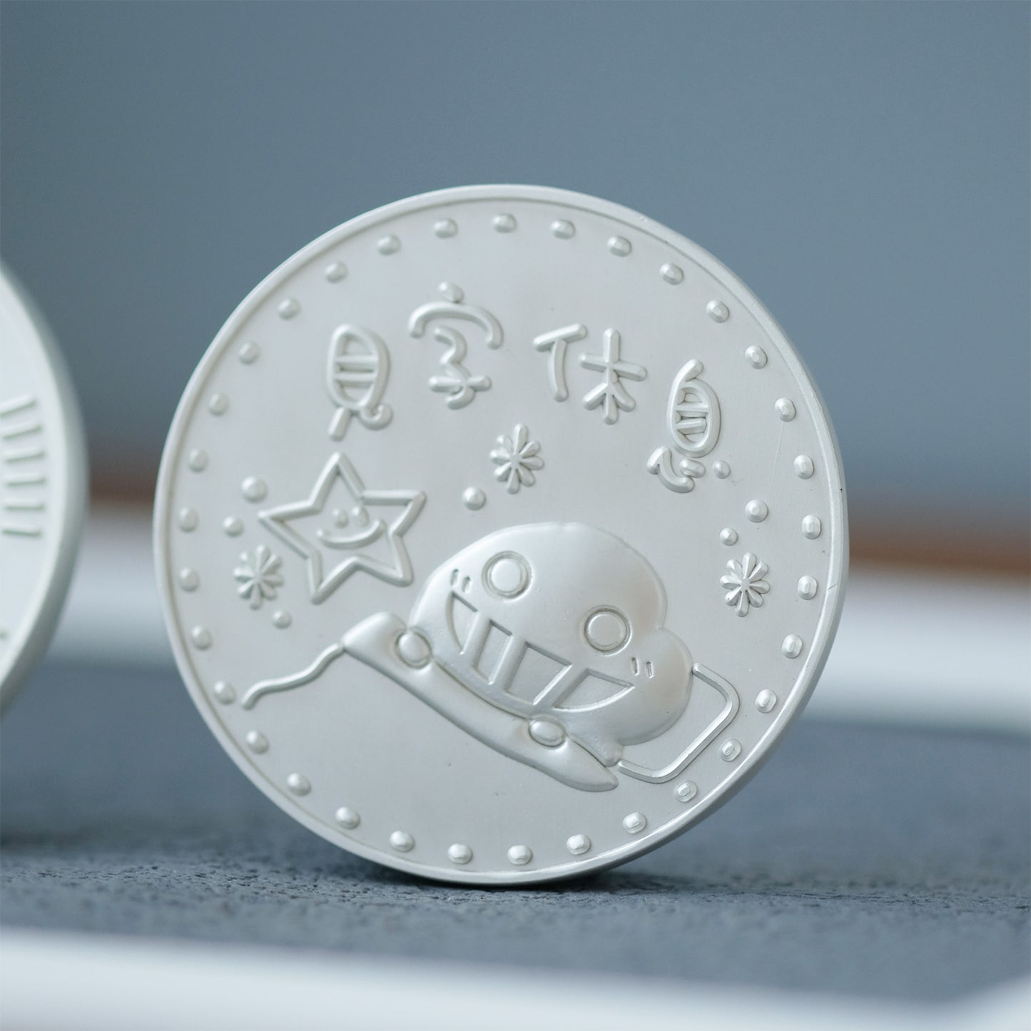 White embossed coin with a design of a robot and stars on a gray surface.